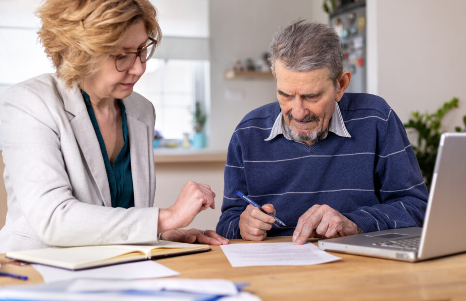Elderly man reviewing and signing documents with assistance from a professional in a home setting, symbolizing personalized power of attorney services